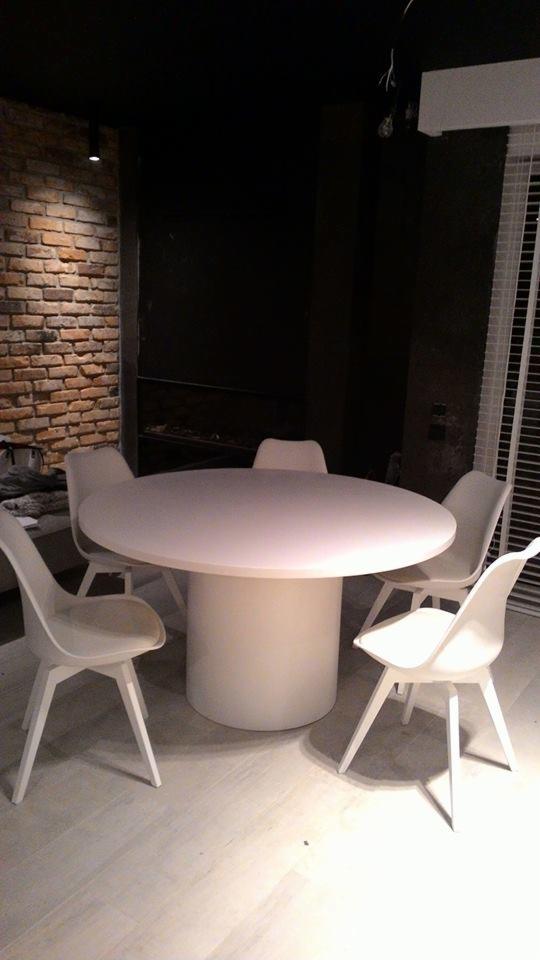 Okrągły, biały stół na cylindrycznej podstawie otoczony czterema białymi krzesłami w minimalistycznym wnętrzu z ceglaną ścianą i ciemnym tłem.