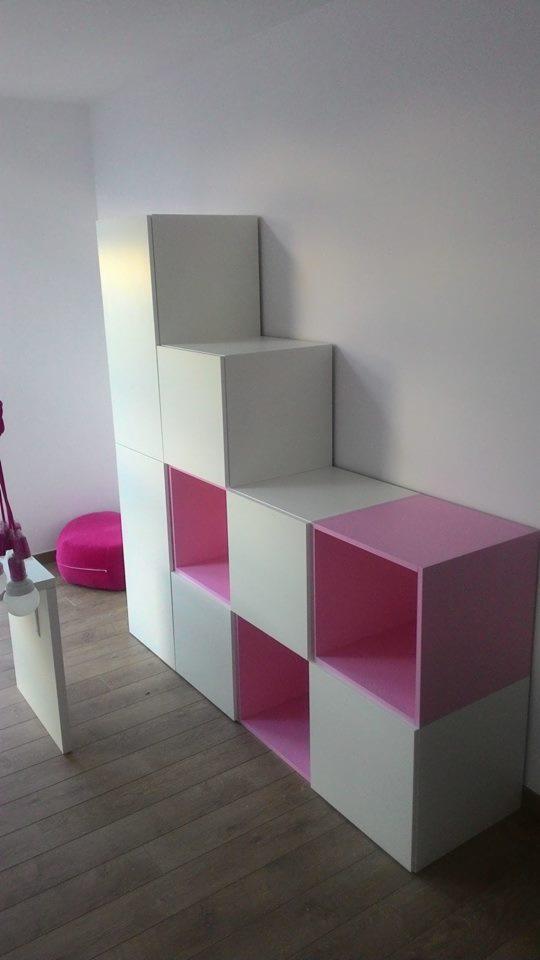 Biało-różowy regał modułowy z otwartymi półkami, ułożony schodkowo, na drewnianej podłodze, z różową pufą w tle.