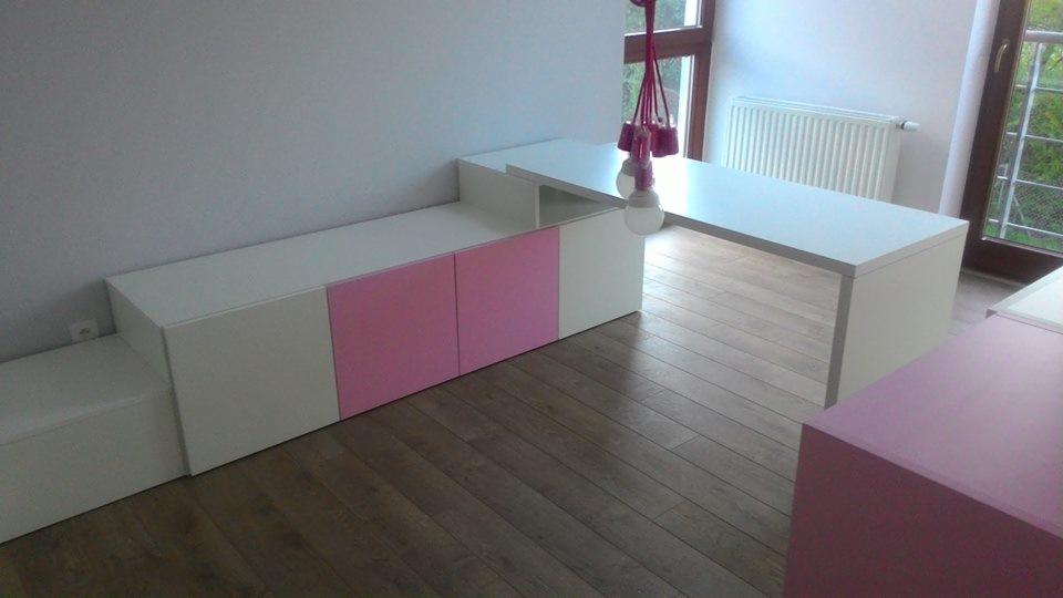 Biało-różowy zestaw mebli na wymiar, obejmujący szafkę RTV i biurko, na drewnianej podłodze z widocznym grzejnikiem i lampą wiszącą.