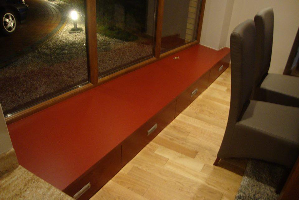 Czerwony, zabudowany parapet z szufladami, drewniana rama okienna z widokiem na podjazd i lampę oświetleniową, parkiet i krzesła w tle.