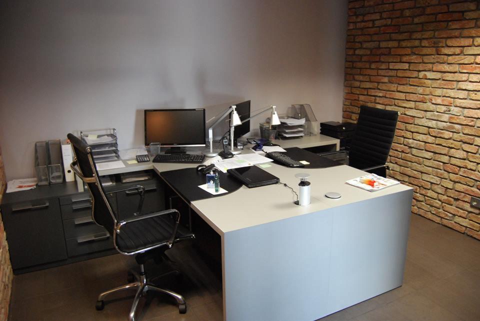 Biuro z biurkiem narożnym w odcieniach szarości, z krzesłem obrotowym i ścianą z cegły w tle.