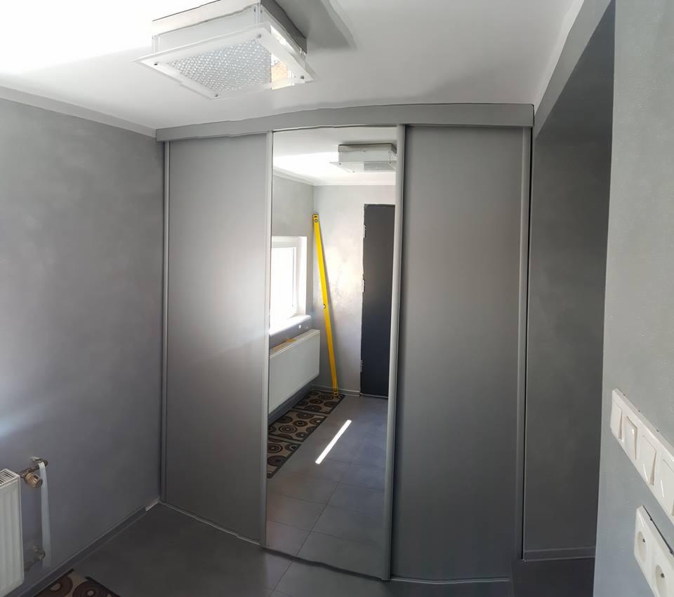 Szara szafa przesuwna z lustrem w przedpokoju, widoczna poziomica oparta o ścianę w głębi, szare ściany i sufit z kwadratową lampą.