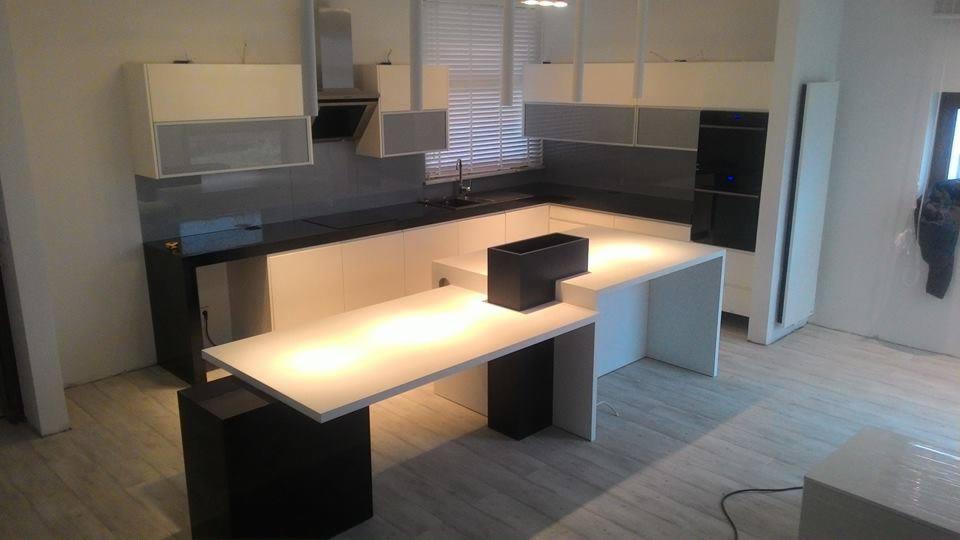 Nowoczesna kuchnia z białymi szafkami i czarnym blatem, wyspa z przedłużonym blatem podświetlonym od spodu, czarny donica na wyspie, szare płytki na ścianie, w tle okno z żaluzjami.