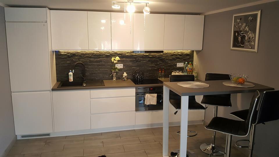 Nowoczesna, biała kuchnia z blatem w kolorze szarości i kamienną ścianą nad blatem, uzupełniona stołem barowym i krzesłami w tle obraz na ścianie.