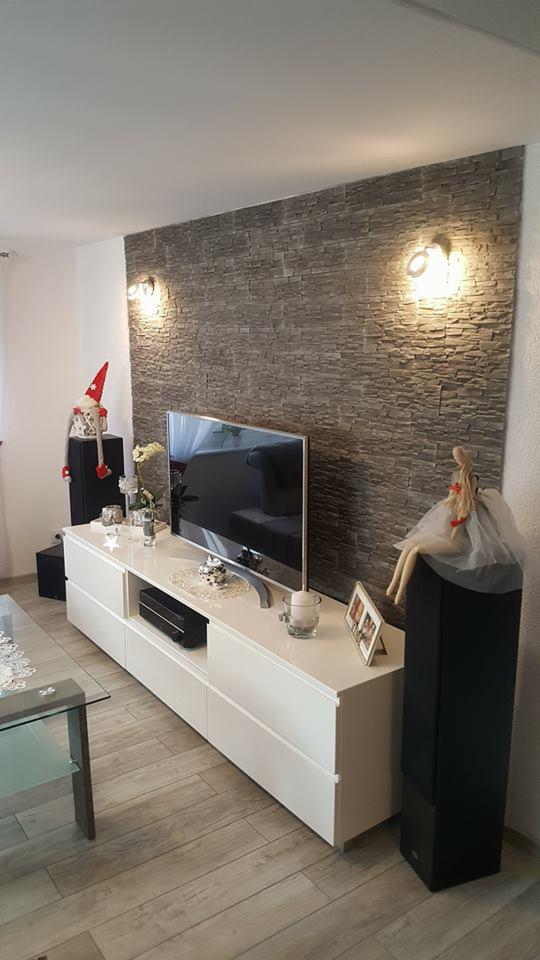 Biała szafka RTV z dekoracjami, na tle ściany wyłożonej szarym kamieniem dekoracyjnym i dwóch kinkietów, w nowoczesnym salonie.