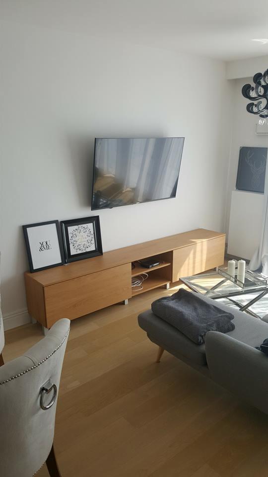 Nowoczesna szafka RTV z jasnego drewna z otwartą półką, wiszący telewizor, dekoracyjne ramki i szary narożnik w minimalistycznym salonie.