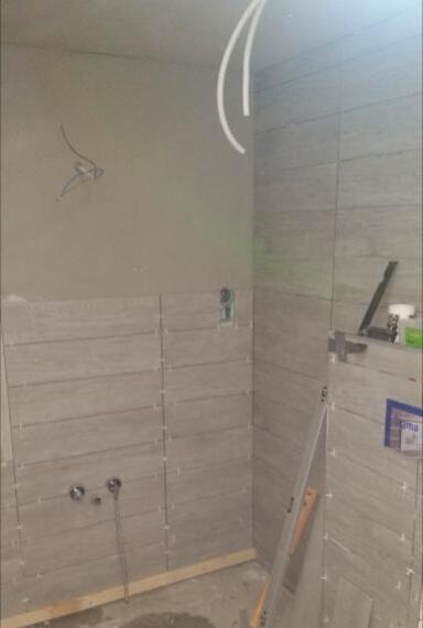 Układanie szarych płytek imitujących drewno w łazience, widoczne krzyżyki dystansowe, niezakończone prace, wystające kable i rury, poziomica oparta o ścianę.