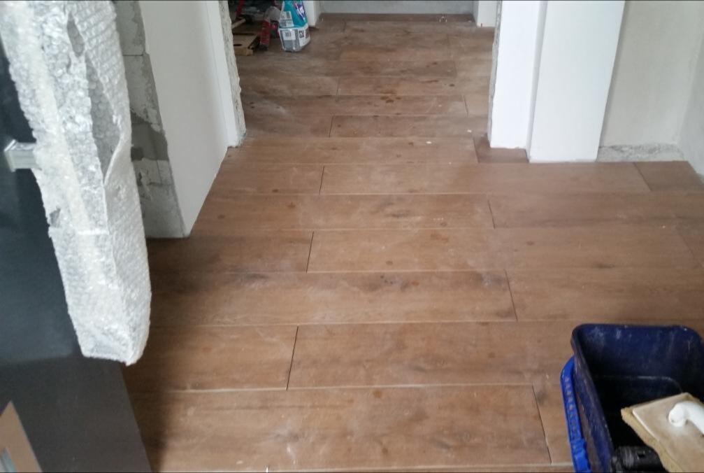 Ułożone płytki podłogowe imitujące drewno, widoczny fragment drzwi w folii ochronnej oraz wiadro z akcesoriami do fugowania.