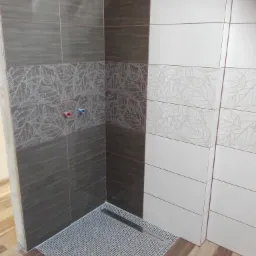 Fragment łazienki z ułożonymi płytkami w odcieniach brązu i bieli, z dekoracyjnym pasem z motywem liści, widoczny odpływ liniowy w strefie prysznicowej i fragment podłogi z imitacją drewna.