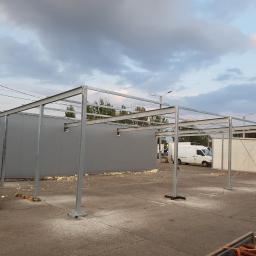 Sob-Stal - Montaż szkieletu stalowego z profili zamkniętych na betonowej posadzce, widoczny budynek z szarej blachy trapezowej w tle i biały samochód dostawczy, pochmurne niebo.