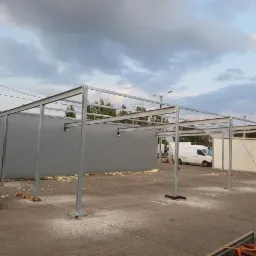 Montaż szkieletu stalowego z profili zamkniętych na betonowej posadzce, widoczny budynek z szarej blachy trapezowej w tle i biały samochód dostawczy, pochmurne niebo.