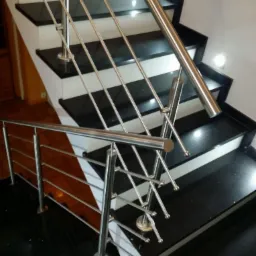 Nowoczesne schody z czarnymi stopniami i białymi podstopnicami, zabezpieczone balustradą ze stali nierdzewnej z poziomymi prętami, widoczne oświetlenie punktowe.