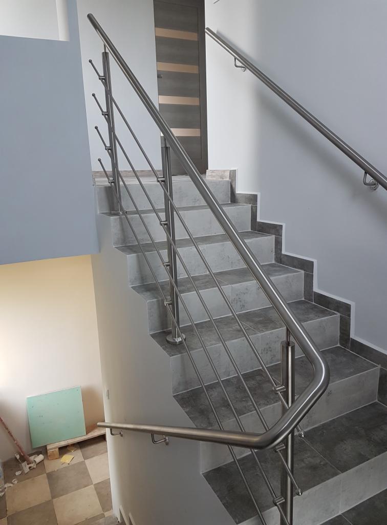 Nowoczesne schody z szarymi stopniami i balustradą ze stali nierdzewnej, widok z dołu na górę, fragment podłogi w szachownicę i drzwi w tle.