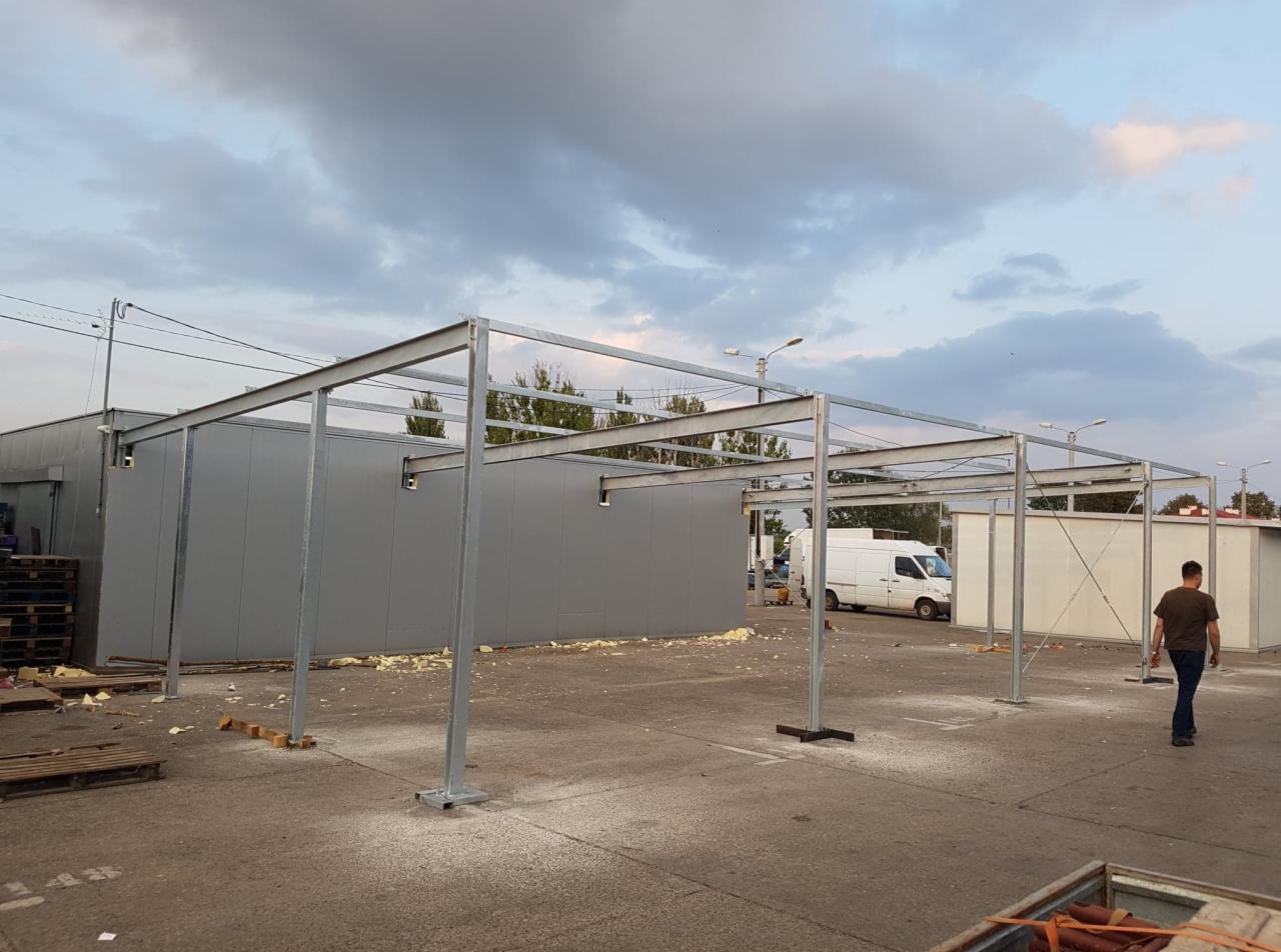 Montaż szkieletu stalowego z profili zamkniętych na betonowej posadzce, widoczny budynek z szarej blachy trapezowej w tle i biały samochód dostawczy, pochmurne niebo.