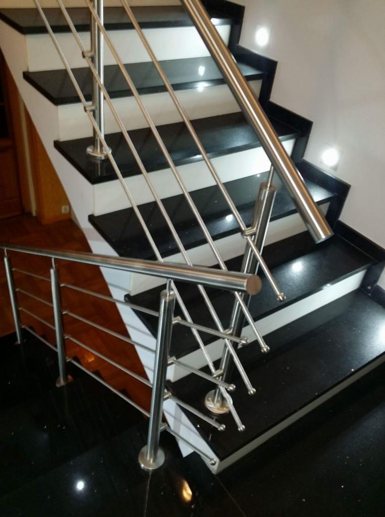 Nowoczesne schody z czarnymi stopniami i białymi podstopnicami, zabezpieczone balustradą ze stali nierdzewnej z poziomymi prętami, widoczne oświetlenie punktowe.