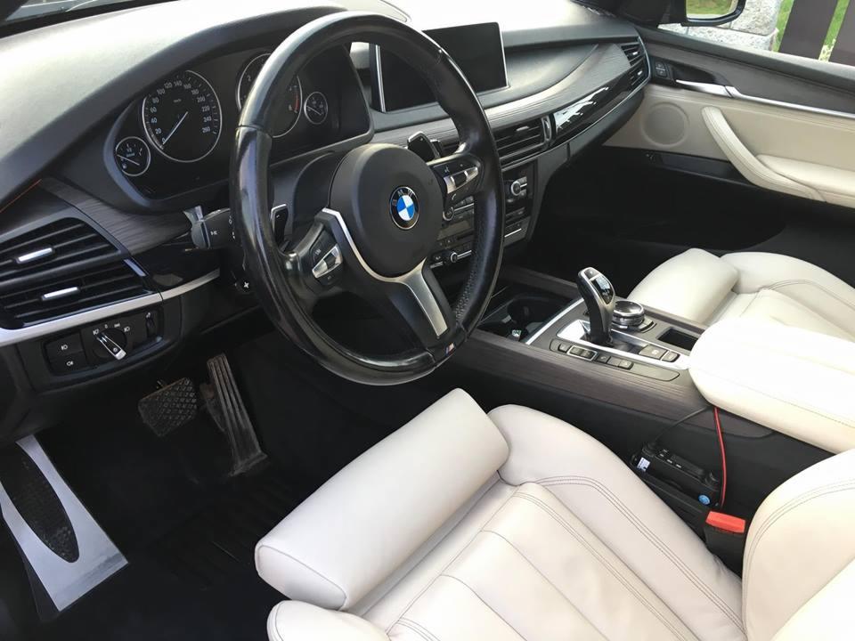 Wnętrze samochodu BMW X5 z jasną skórzaną tapicerką, widok z boku na deskę rozdzielczą, kierownicę z logo BMW i fotel pasażera.