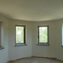 Wnętrze okrągłego pomieszczenia z czterema oknami po wykonaniu białych gładzi na ścianach i suficie, widoczne zabezpieczenia folią okien i parapetów.