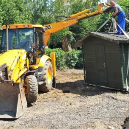 Ż&oacute;łta koparko-ładowarka podnosi gotową, drewnianą altanę, kt&oacute;rą zabezpiecza mężczyzna w niebieskim kombinezonie stojący na dachu, obok drabina oparta o ścianę, w tle zieleń.