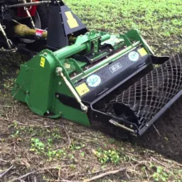 Tractor with green soil tiller attachment, preparing the ground for planting, visible tilled soil and remaining grass patches.