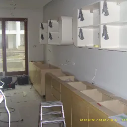 Montaż kuchni na wymiar w trakcie realizacji: dolne szafki bez blatów, wiszące szafki z uchwytami w kształcie litery A, drabina i narzędzia na podłodze.