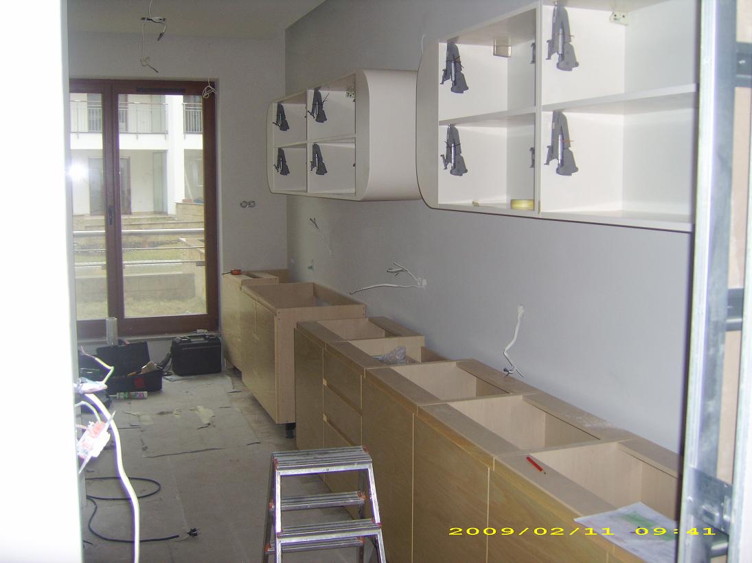 Montaż kuchni na wymiar w trakcie realizacji: dolne szafki bez blatów, wiszące szafki z uchwytami w kształcie litery A, drabina i narzędzia na podłodze.