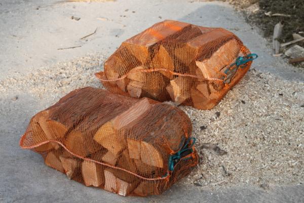 Dwa worki siatkowe wypełnione pociętym drewnem ułożone na betonowej powierzchni, otoczone drobnymi wiórami.