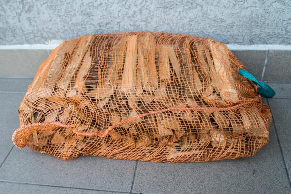 Worek z rżniętym drewnem opałowym, widoczna siatka w kolorze pomarańczowym i zielony uchwyt, tło stanowi szara ściana i płytki.