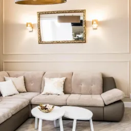 Beżowa narożna sofa z dekoracyjnymi poduszkami w salonie z kremowymi ścianami, złotym lustrem i dwoma małymi białymi stolikami kawowymi.