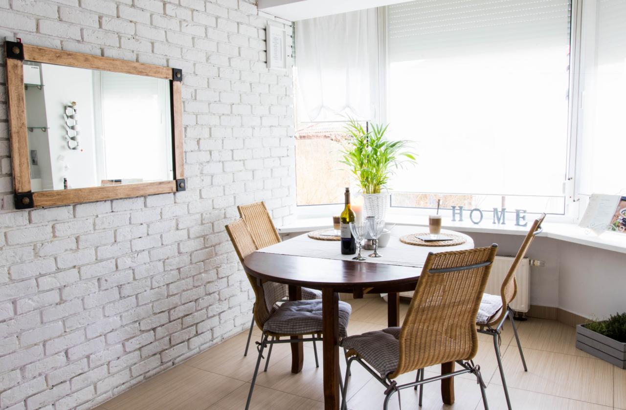 Jasna jadalnia z okrągłym drewnianym stołem, wiklinowymi krzesłami z szarymi poduszkami, białymi ceglanymi ścianami, lustrem w drewnianej ramie i rośliną w doniczce na parapecie.