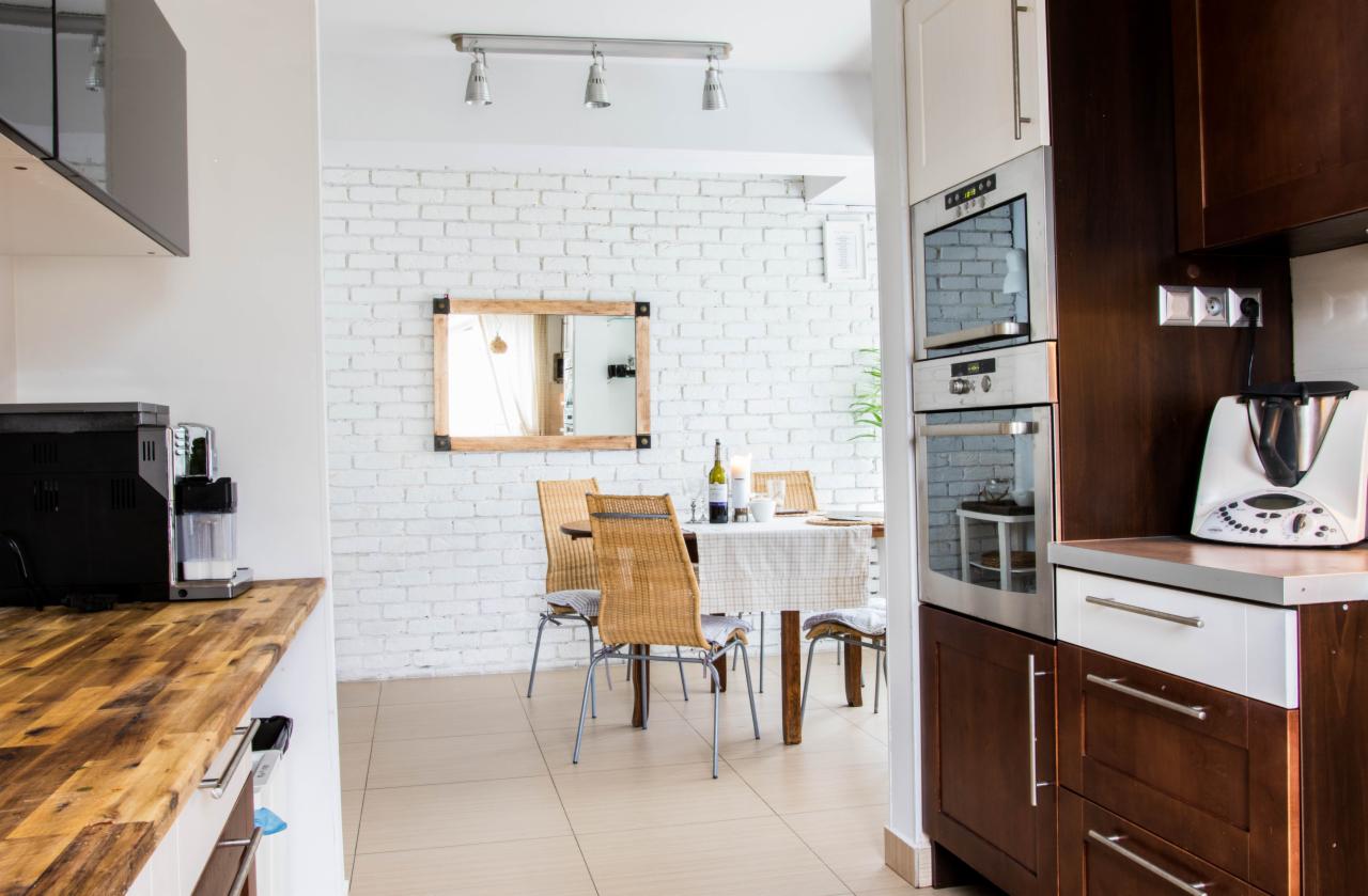 Jasne wnętrze kuchni z jadalnią, biała cegła na ścianie, stół z wiklinowymi krzesłami, drewniany blat i nowoczesny ekspres do kawy.