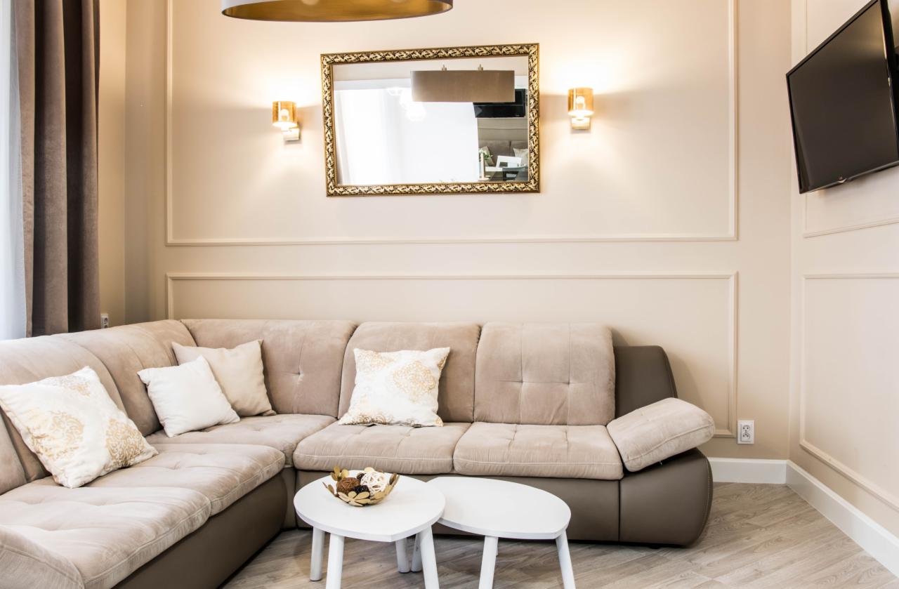 Beżowa narożna sofa z dekoracyjnymi poduszkami w salonie z kremowymi ścianami, złotym lustrem i dwoma małymi białymi stolikami kawowymi.