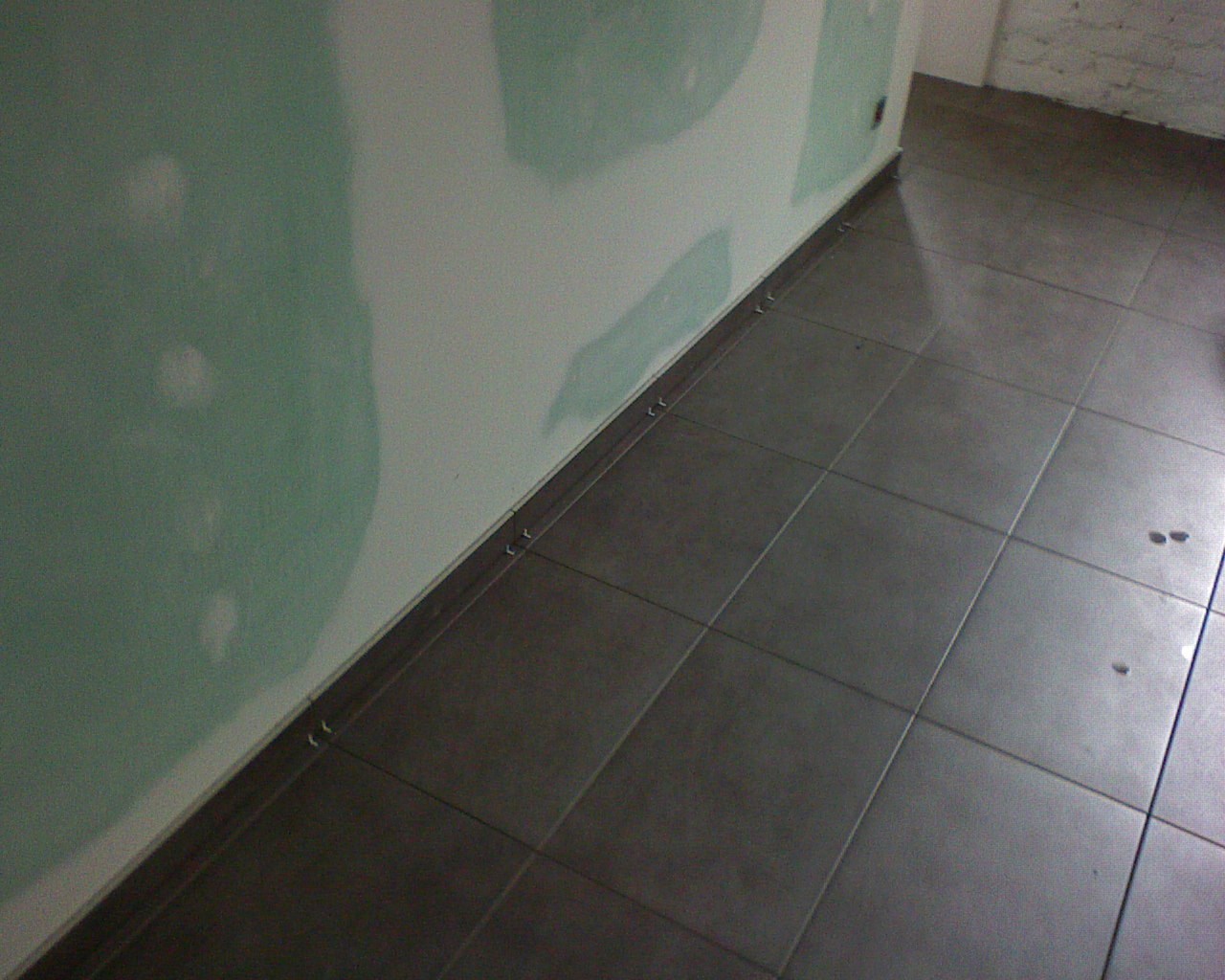 Ułożone ciemnoszare płytki podłogowe przy ścianie z płyt gipsowo-kartonowych, widoczne metalowe listwy przypodłogowe, fragment białej ściany w tle.