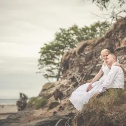 Para młoda w białych strojach siedzi na klifie z widokiem na morze podczas sesji wideofilmowania ślubnego, bukiet leży obok nich.