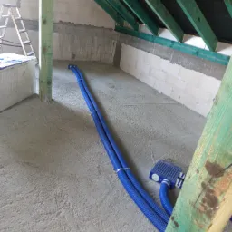 Montaż rekuperacji na poddaszu: Dwa niebieskie przewody wentylacyjne ułożone na szarej posadzce, widoczne drewniane elementy konstrukcyjne dachu i ściany z betonu komórkowego.