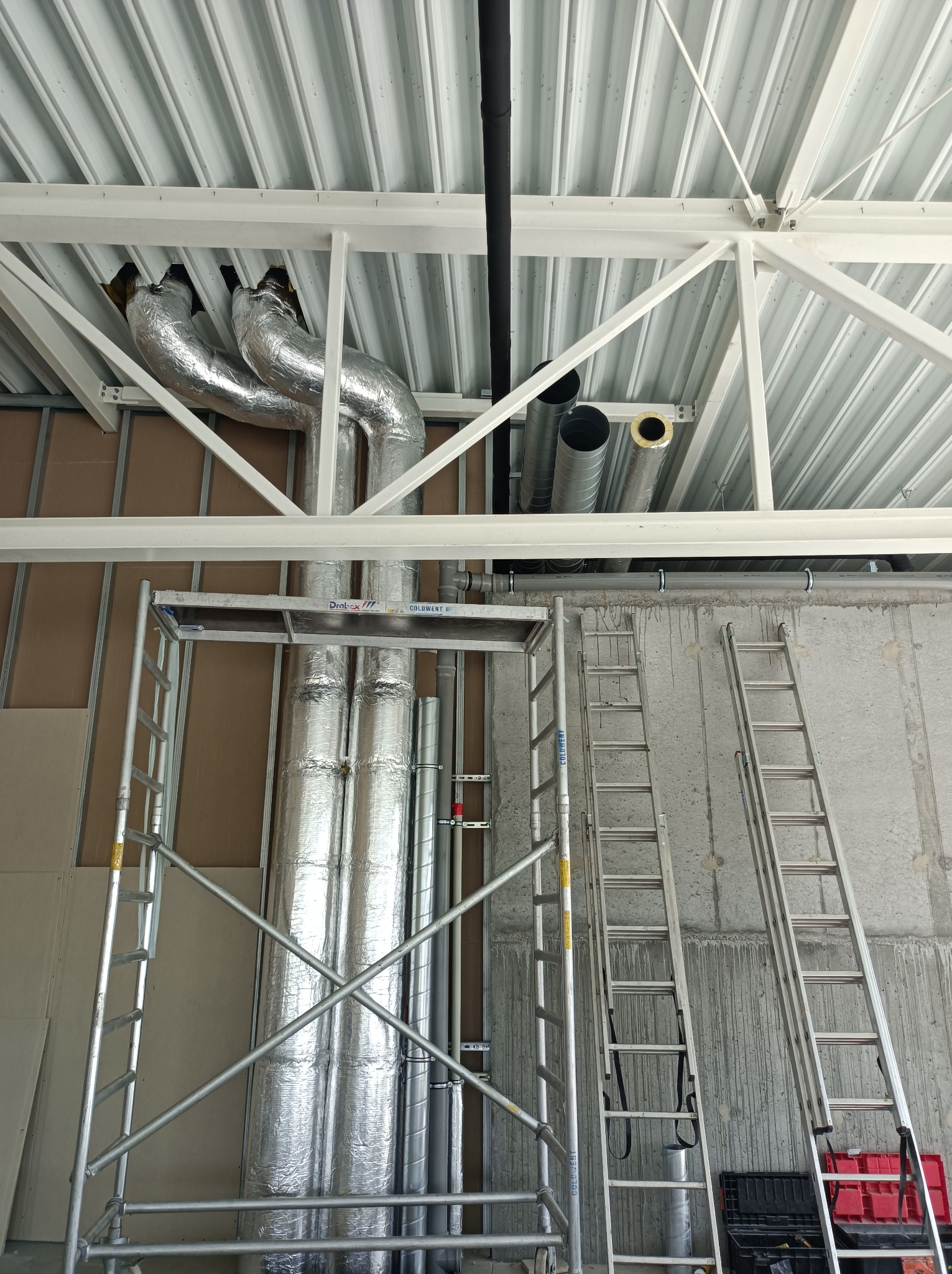 Montaż izolowanych przewodów wentylacyjnych pod stropem z blachy trapezowej. Widoczne rusztowanie i drabiny.