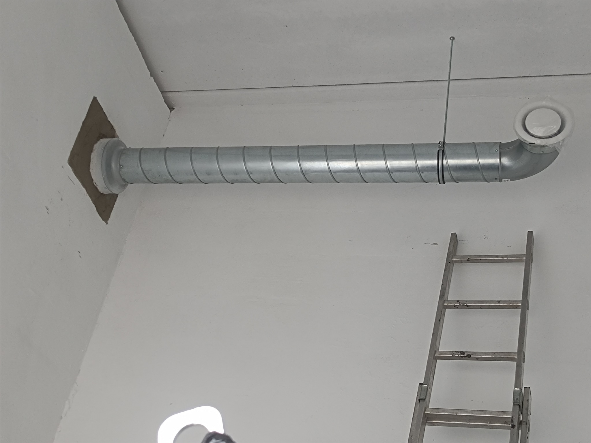 Sztywny kanał wentylacyjny spiralny, zamocowany do sufitu, z okrągłym anemostatem. Obok drabina aluminiowa oparta o ścianę. Widok z dołu.