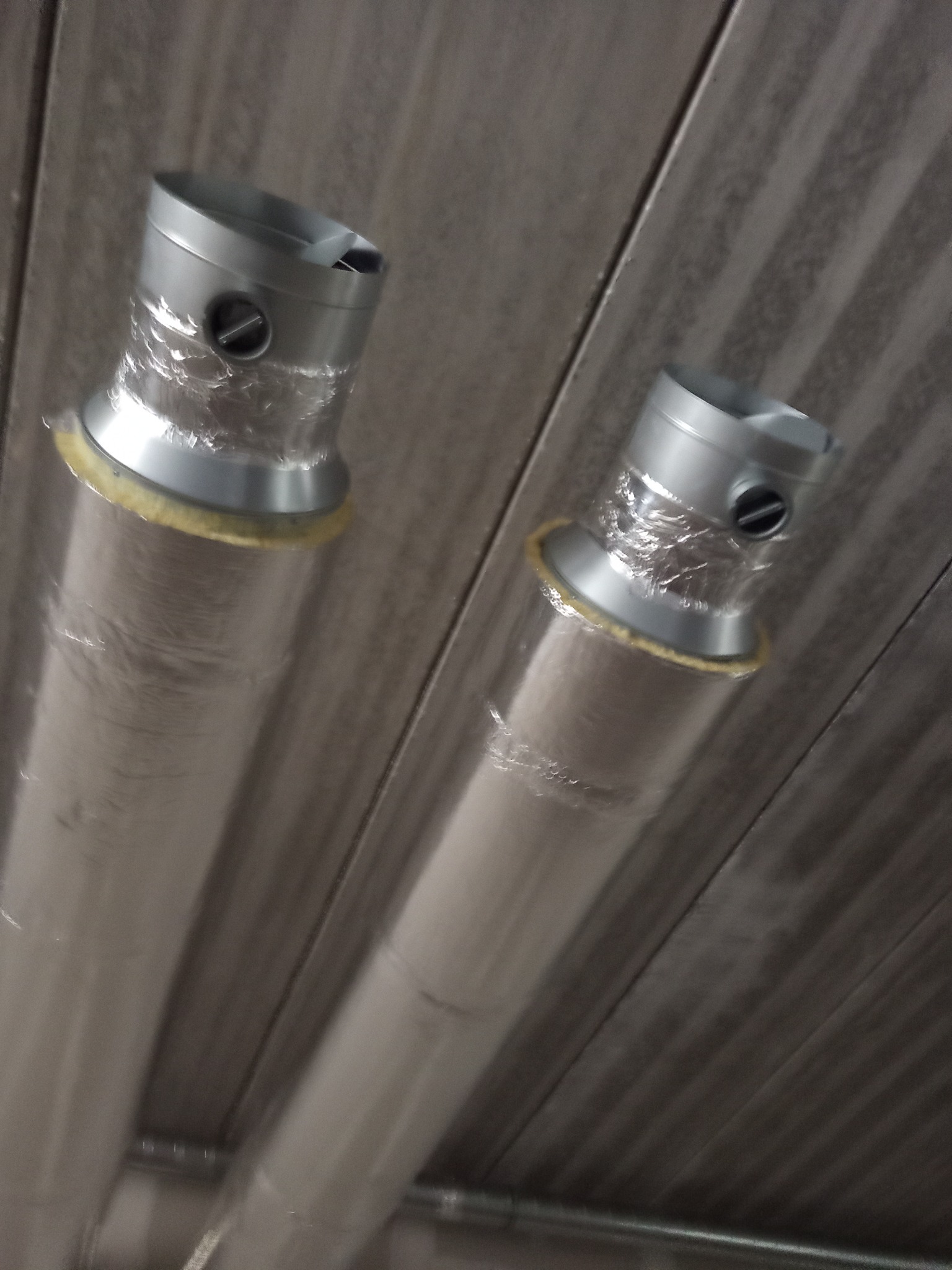 Dwa przewody wentylacyjne z srebrną izolacją, zamontowane pod kątem na szarej, żebrowanej powierzchni. Widoczne elementy mocujące i fragment rury w tle.