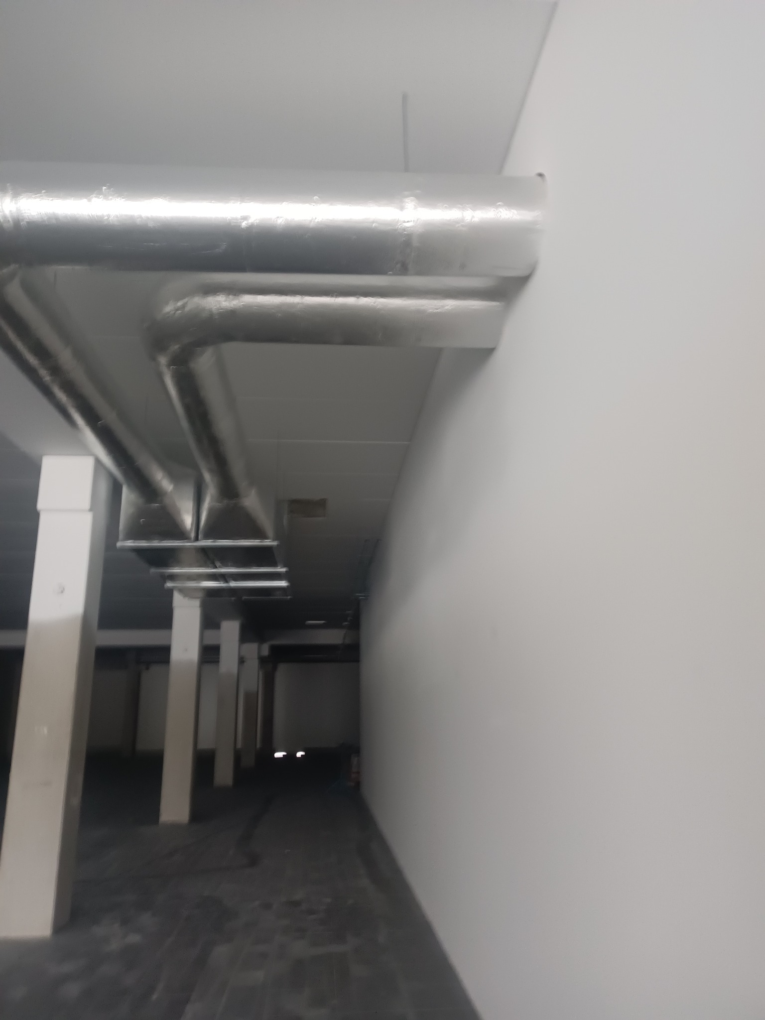 Srebrne rury wentylacyjne zamontowane pod sufitem w hali przemysłowej, widoczne białe filary i szara podłoga. Układ rur równoległy do ściany.