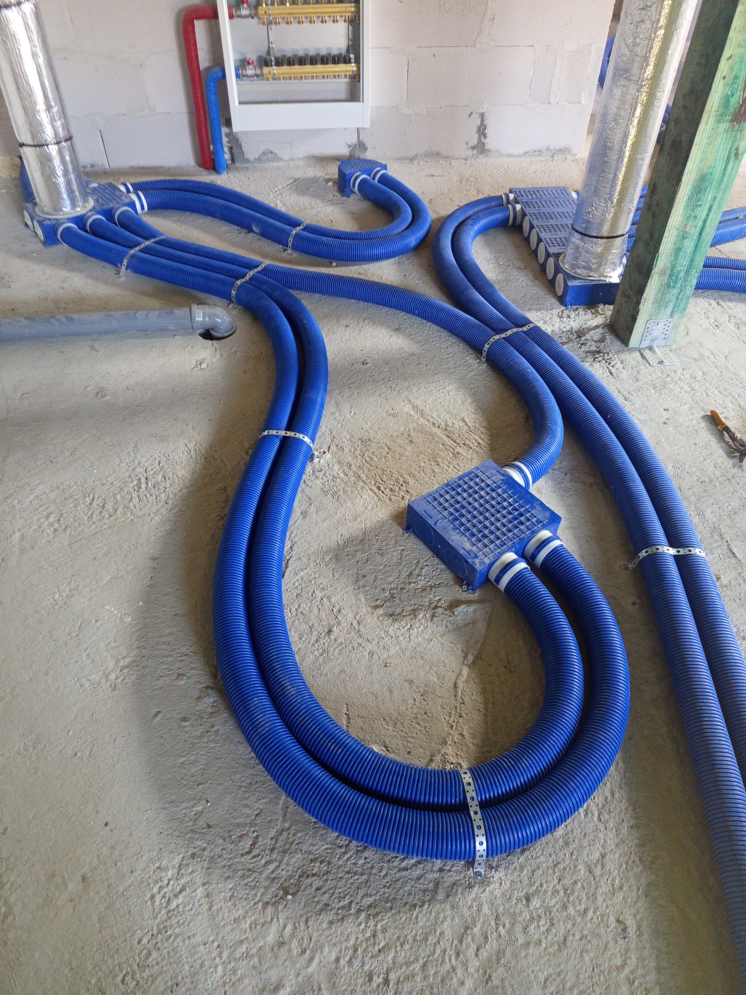 Niebieskie, karbowane rury wentylacyjne ułożone na surowym podłożu, połączone w system. Widoczne elementy montażowe i skrzynka rozdzielcza na ścianie w tle.