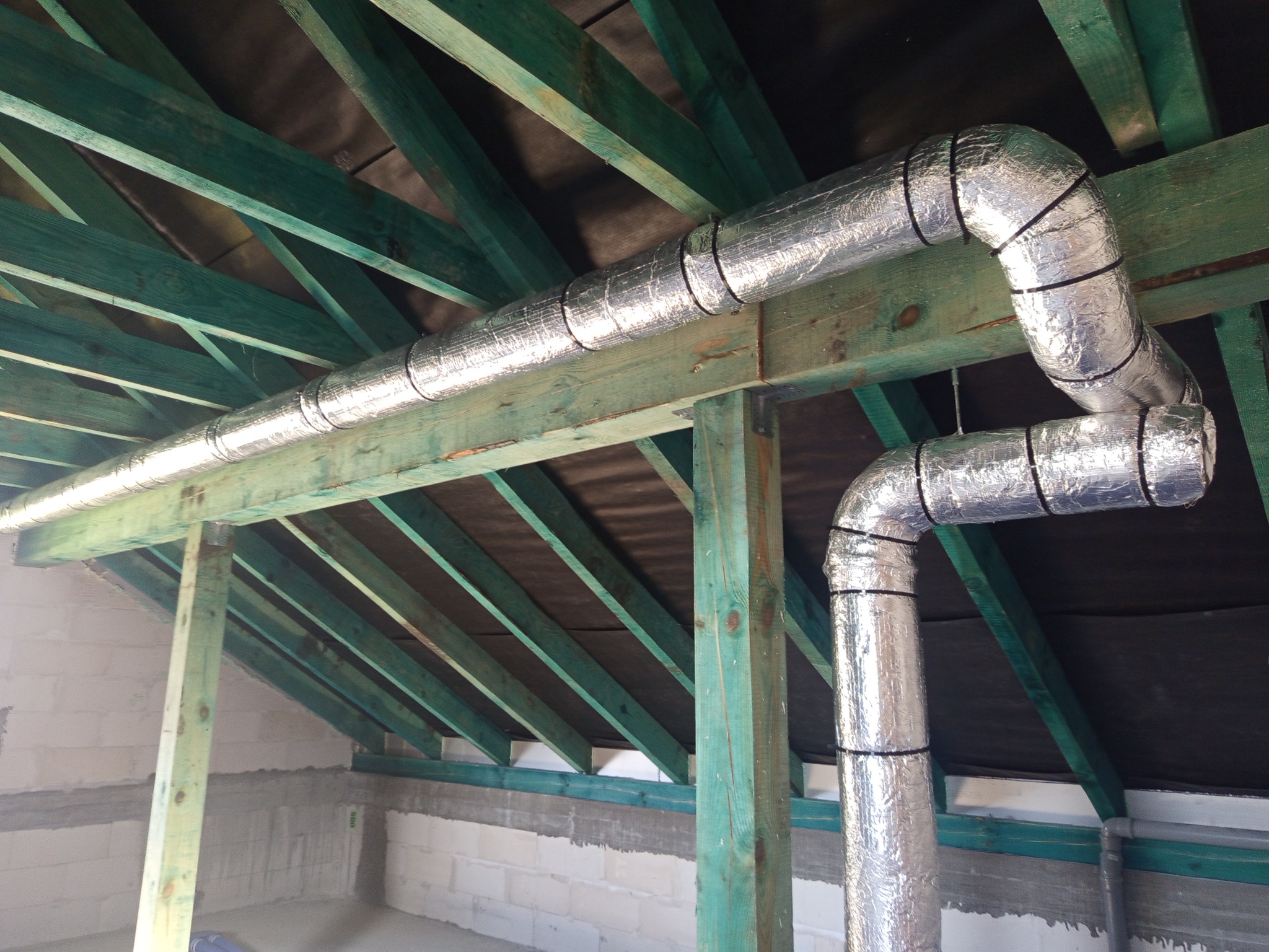 Izolowane przewody wentylacyjne z folii aluminiowej zamontowane pod zielonymi krokwiami dachu na poddaszu w surowym stanie.