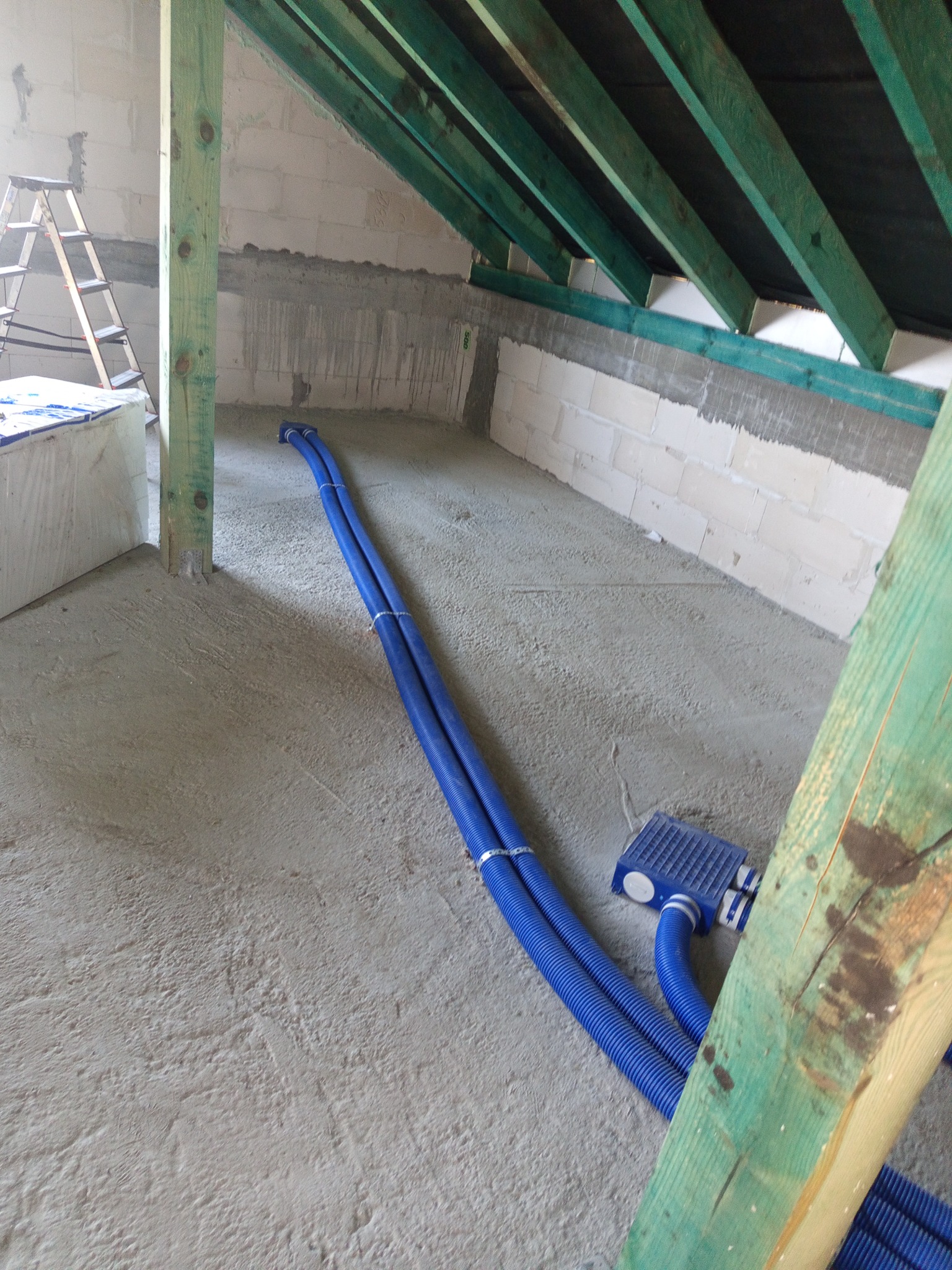 Montaż rekuperacji na poddaszu: Dwa niebieskie przewody wentylacyjne ułożone na szarej posadzce, widoczne drewniane elementy konstrukcyjne dachu i ściany z betonu komórkowego.