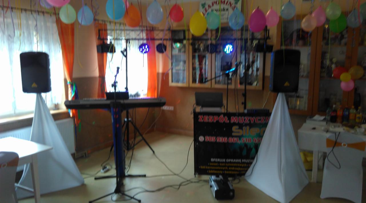 Sprzęt zespołu muzycznego przygotowany do występu w udekorowanym sali z balonami, widoczne kolumny na białych pokrowcach, keyboard, oświetlenie sceniczne i stolik z napojami.