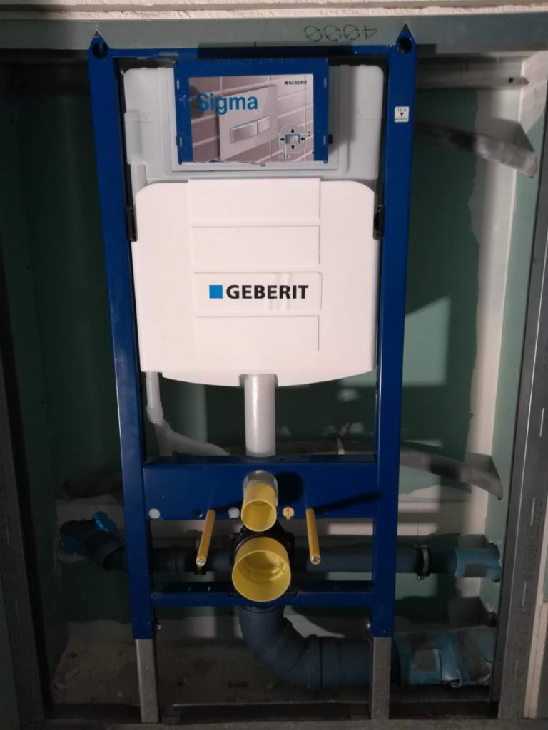 Stelaż podtynkowy Geberit Sigma w trakcie montażu, widoczne niebieskie elementy konstrukcyjne, biały zbiornik i żółte złączki kanalizacyjne w surowym otoczeniu ściany z płyt gipsowo-kartonowych.