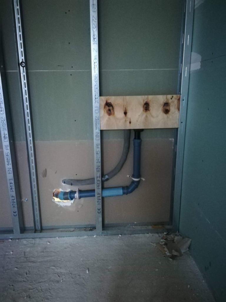 Surowy stan instalacji wodnej w łazience: rury PCV szare i niebieskie podłączone do ściany z płyt gipsowo-kartonowych, wsparte na drewnianej belce z otworami, widoczne profile metalowe konstrukcji...