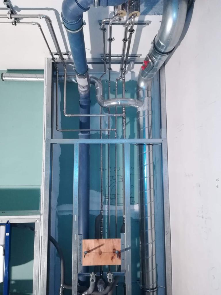 Widok na instalację rur różnego typu i średnicy w metalowej ramie w trakcie montażu wewnątrz ściany, widoczne elementy mocujące i notatki na ścianie.