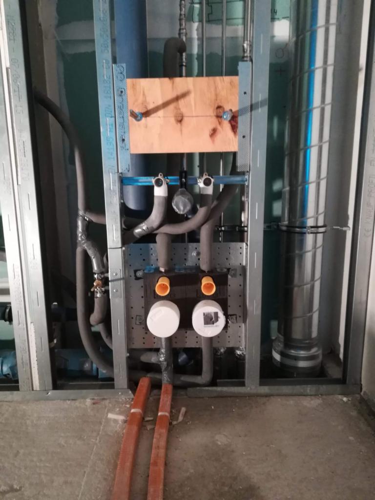 Wewnętrzna instalacja wodno-kanalizacyjna w trakcie montażu w budynku, widoczne rury, zawory, mocowania na metalowej konstrukcji szkieletowej, z fragmentem drewnianej deski montażowej.