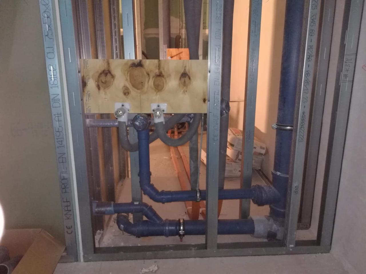 Wewnętrzna instalacja wodno-kanalizacyjna w trakcie montażu, widoczne niebieskie rury z tworzywa sztucznego, zawory i metalowa konstrukcja szkieletowa ścian działowych.