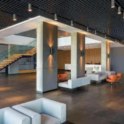 Hotelowe lobby z brązową, kwadratową podłogą z płytek, betonowymi kolumnami z oświetleniem ściennym, drewnianymi schodami ze szklaną balustradą, szarą sofą i pomarańczowymi fotelami oraz recepcją...