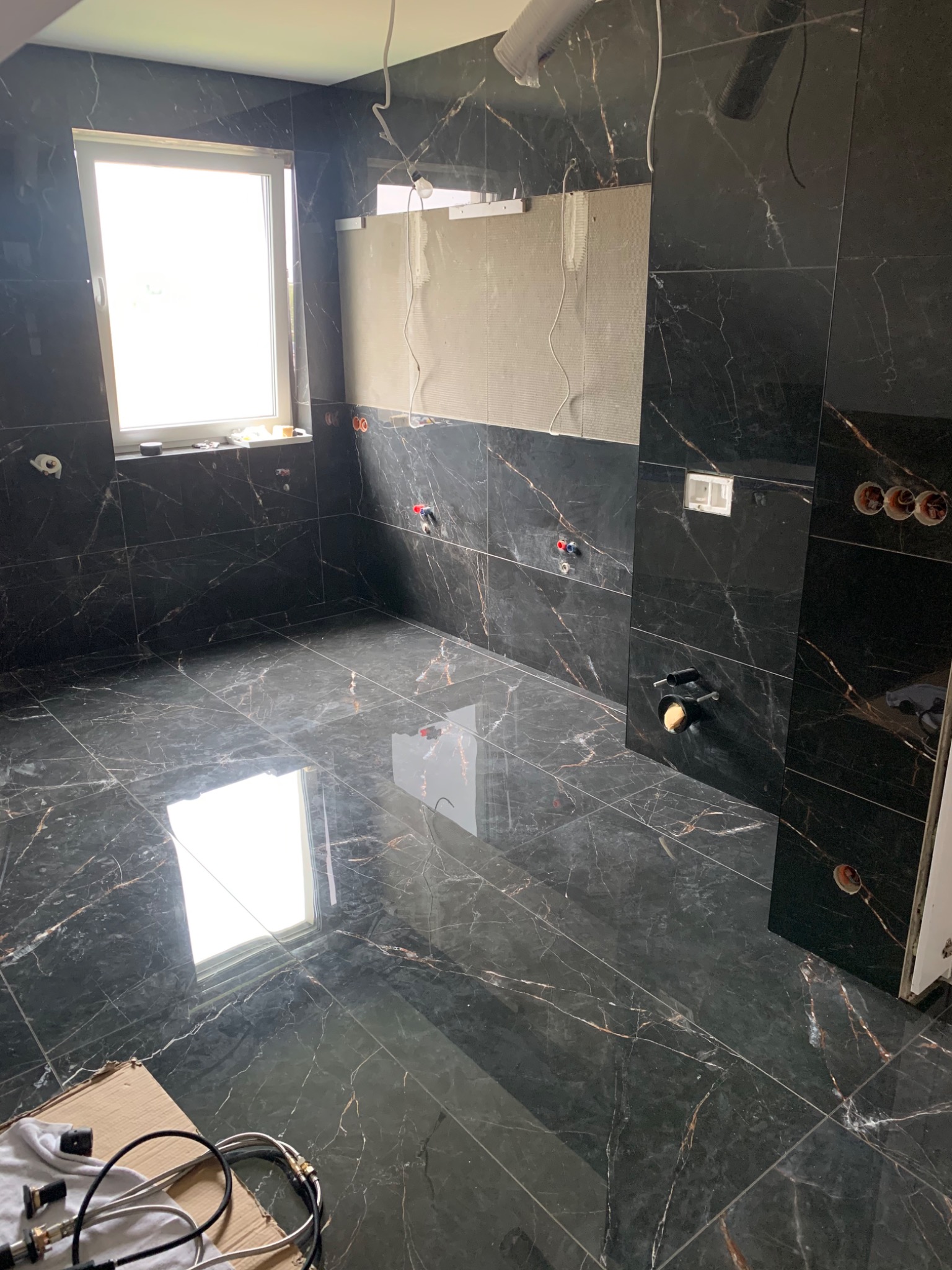 Łazienka w trakcie remontu, wyłożona czarnymi płytkami imitującymi marmur z widocznymi instalacjami hydraulicznymi i fragmentem niezakończonej ściany z płytami gipsowymi.