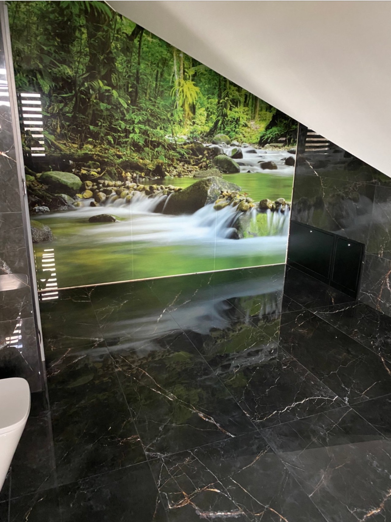 Czarne płytki marmurowe na podłodze i ścianie w łazience z fototapetą przedstawiającą leśny strumień.